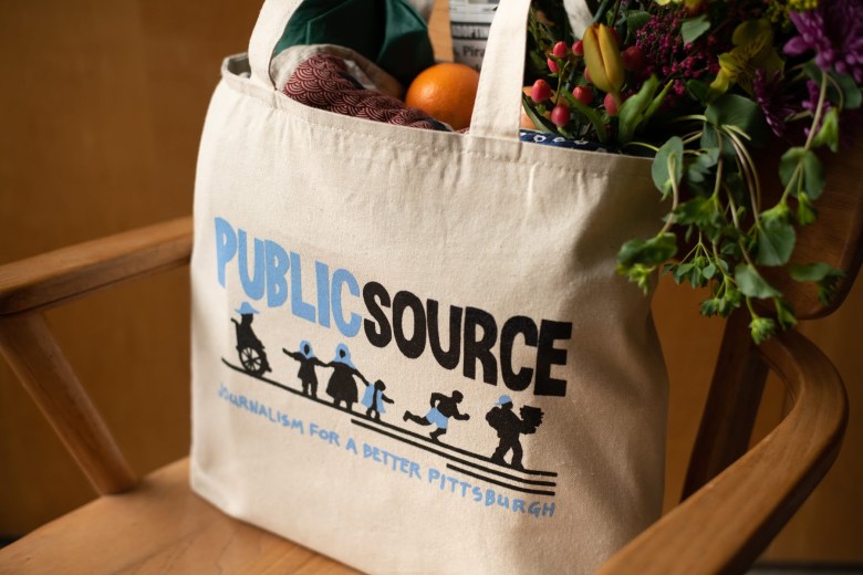 A Publicsource tote bag sitting on a chair, filled with a bouquet of flowers, an umbrella, and fruit.  The tote reads PUBLICSOURCE: Journalism for a better Pittsburgh and shows images of a person in a wheelchair, a person in a headscarf holding the hands of two children, a person running, and a person carrying a stack of books.